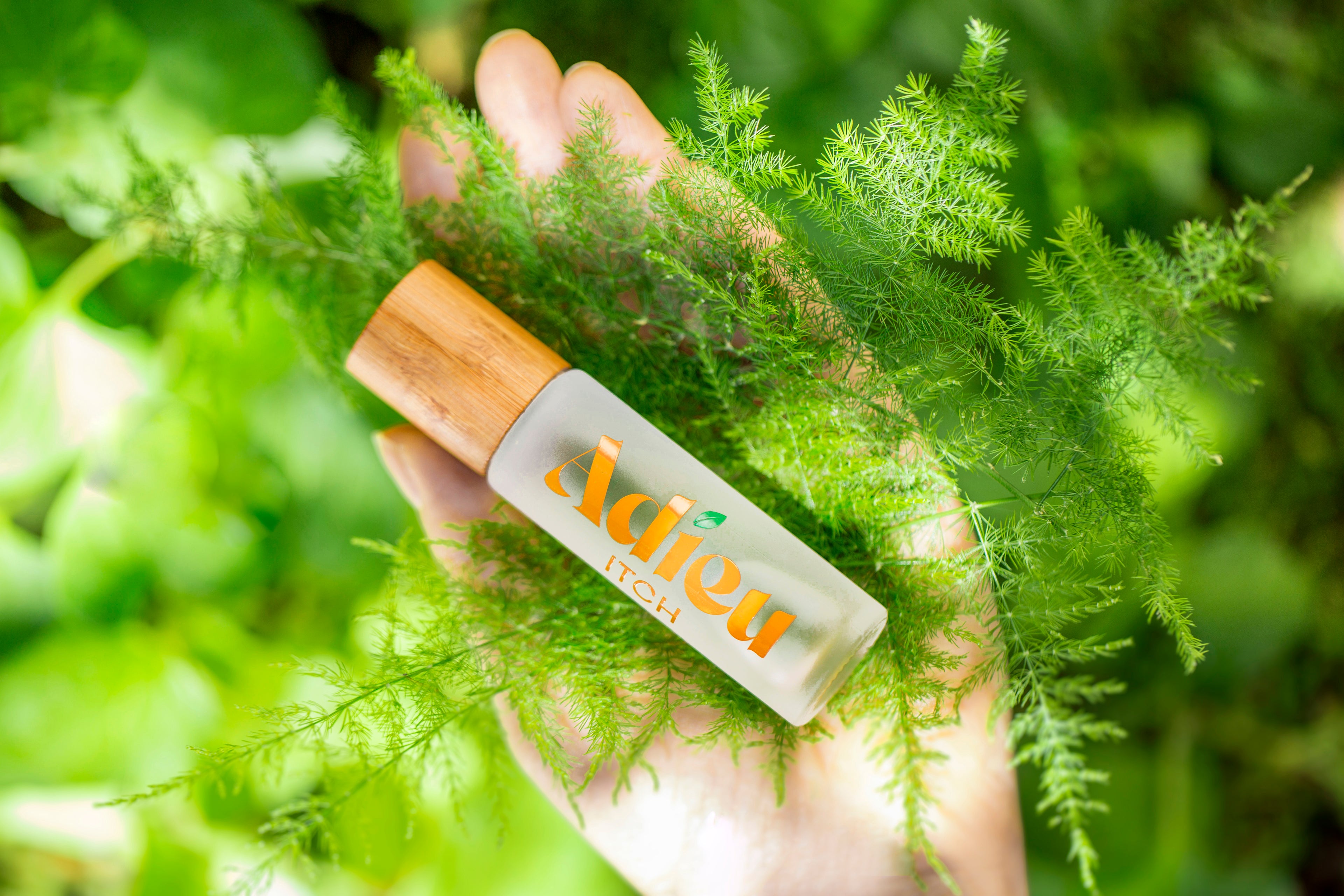 A hand holding a small roll-on bottle with a wooden cap, surrounded by green leafy plants. The bottle has a frosted white body with orange and green 'Adieu NATURE' branding.